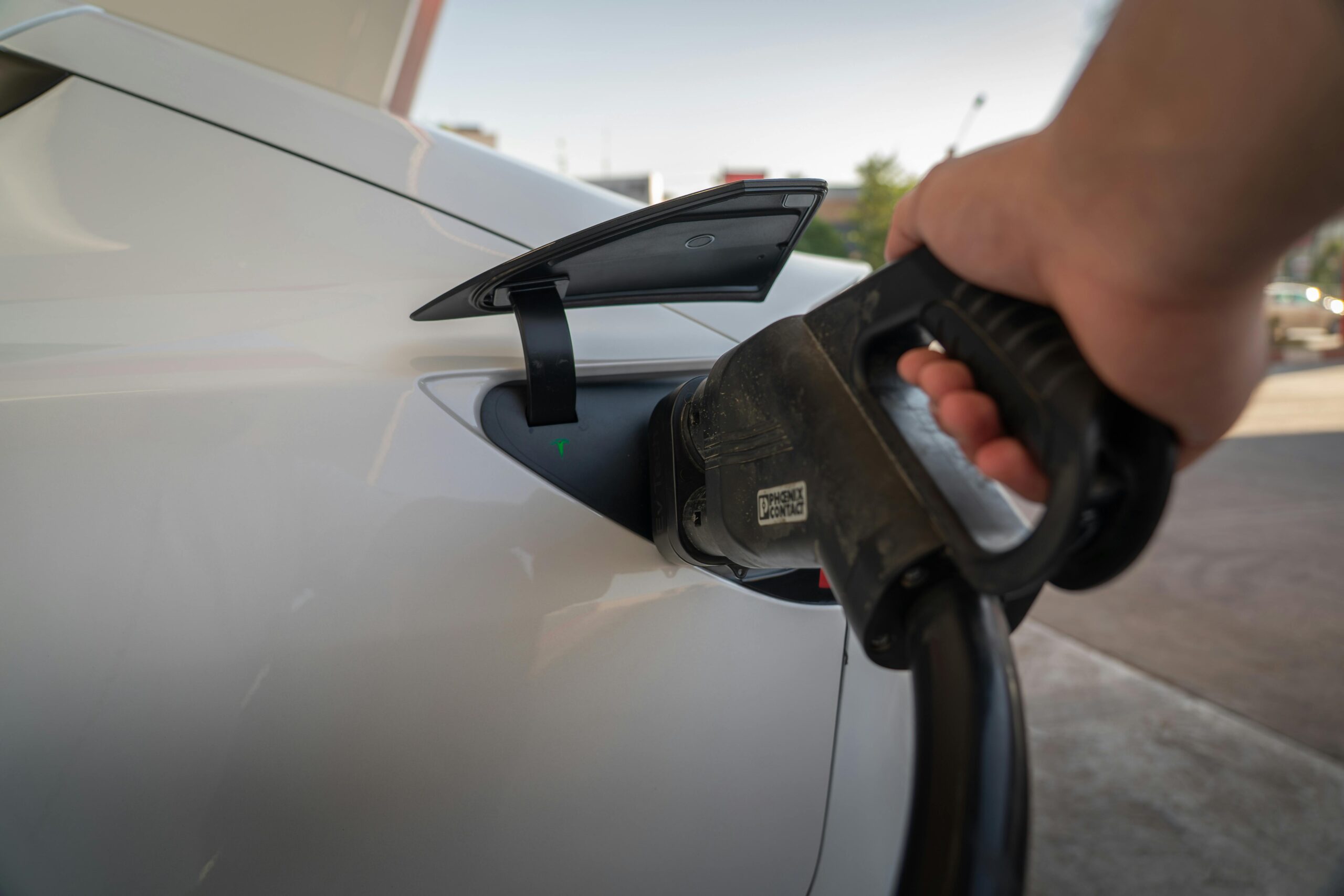 a person’s hand holds a black charging handle plugged into the rear charging port of a white Tesla, with a small green indicator light visible inside the port.