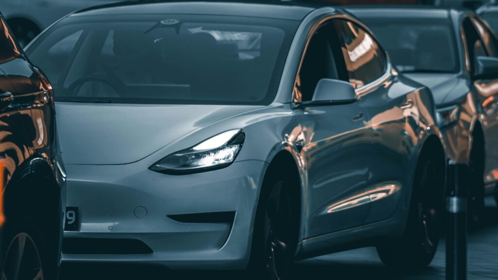 white tesla model 3 parked in a row of cars, with its headlight illuminated and a soft orange reflection on the side panel.